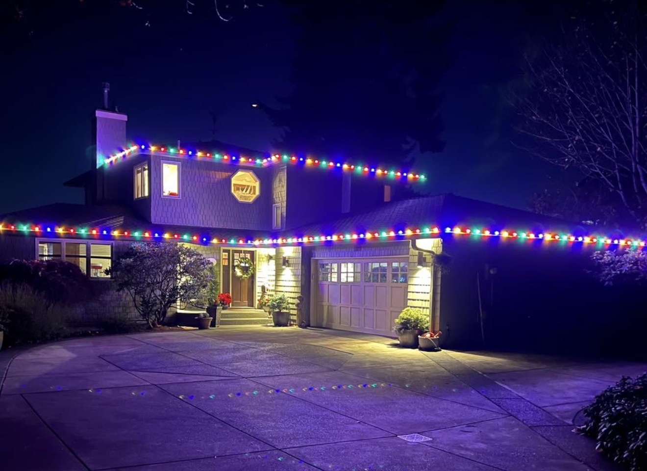 Two-story Kitsap County home professionally lit with warm-white Christmas roofline lights and tree wraps